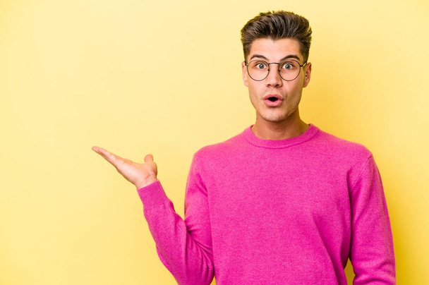 Young caucasian man isolated on yellow background impressed holding copy space on palm. - Foto, immagini