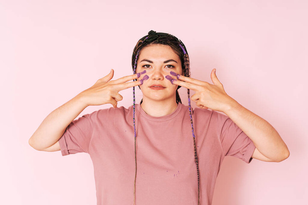 Mädchen malt mit Fingern lila Farbe auf ihr Gesicht vor rosa Hintergrund. - Foto, Bild