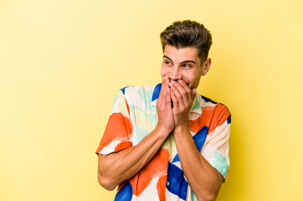 Jonge blanke man geïsoleerd op gele achtergrond lachen over iets, bedekken mond met handen. - Foto, afbeelding