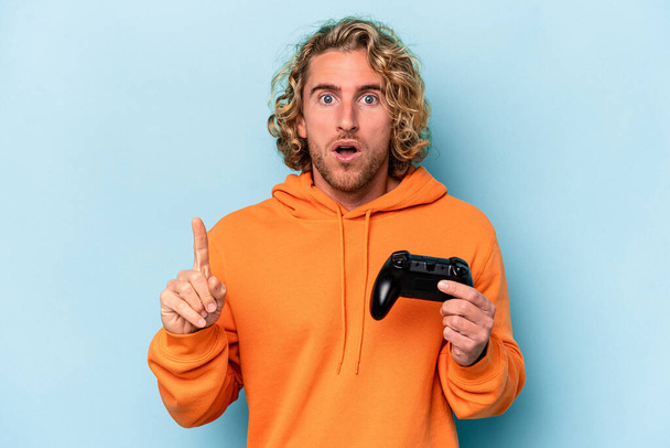 Young caucasian man playing with a video game controller isolated on blue background having some great idea, concept of creativity. - Foto, afbeelding