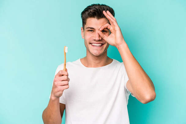 Hombre joven caucásico cepillarse los dientes aislados sobre fondo blanco excitado mantener el gesto bien en el ojo. - Foto, Imagen