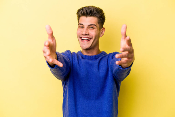 Young caucasian man isolated on yellow background feels confident giving a hug to the camera. - Zdjęcie, obraz