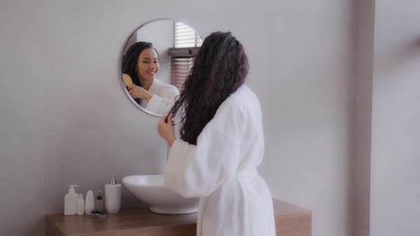 Young happy hispanic woman stands in bathroom in bathrobe looks to mirror combs thick long healthy curly hair using hairbrush feels satisfied morning daily routine advertise haircare treatment concept - Filmati, video