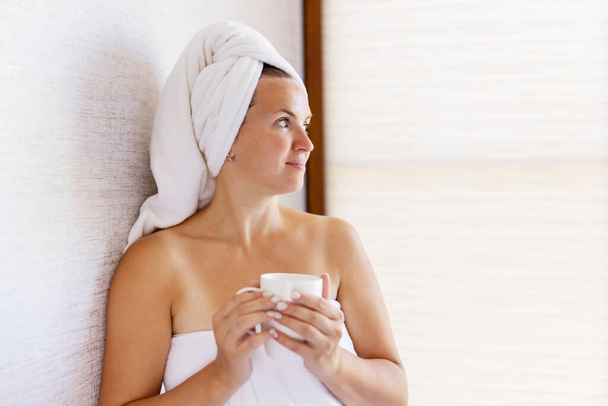 Bella giovane donna con un asciugamano in testa tiene una tazza di caffè al mattino. Concetto di stile di vita domestico. - Foto, immagini