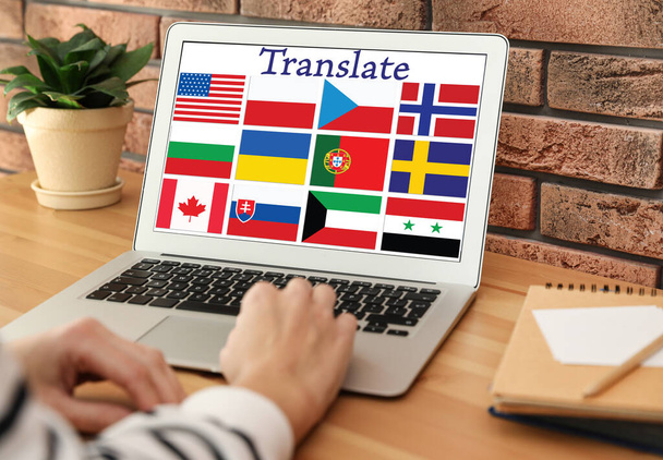 Translator using modern laptop with images of different flags on screen at table indoors, closeup - Фото, зображення