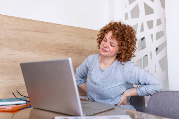 Portret van een jonge gestresste vrouw die thuis op kantoor achter de laptop zit, pijn in de nek met pijnlijke uitdrukking, pijn in de nek na het werken aan de pc - Foto, afbeelding