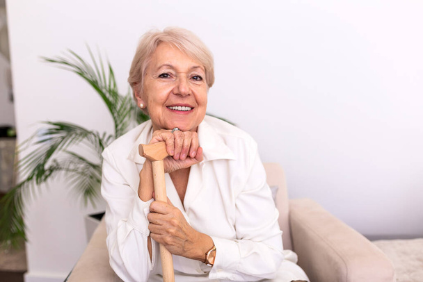 Portrait of beautiful senior woman with white hair and walking stick. Portrait of senior woman sitting on sofa at home. Smiling middle aged mature grey haired woman looking at camera - 写真・画像