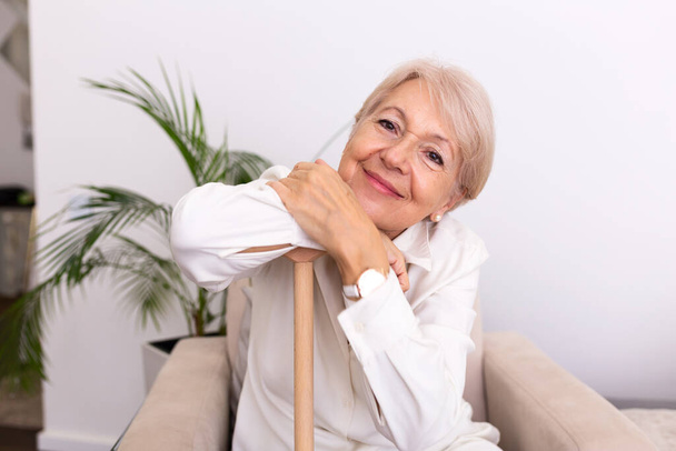 Grand-mère souriante assise sur le canapé. Portrait d'une belle femme âgée souriante avec canne à pied sur fond clair à la maison. Vieille femme assise avec ses mains sur une canne - Photo, image