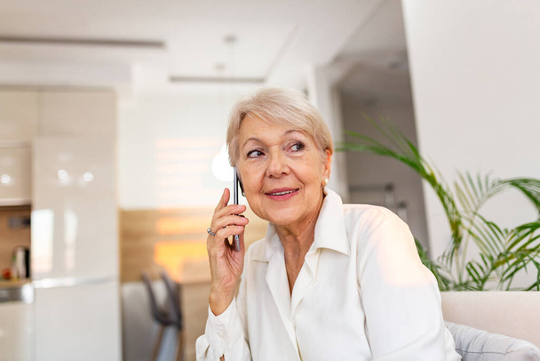 Bella donna anziana caucasica che ha una conversazione telefonica con la sua vecchia amica usando un dispositivo elettronico, discutendo le ultime notizie con un sorriso carino sul suo viso, seduta a casa - Foto, immagini
