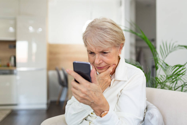 Verwirrte Seniorin hat Probleme mit dem Handy zu Hause. Alte Frau mit weißem Haar sitzt auf Sofa und versucht mit Smartphone Nachrichten zu verschicken. - Foto, Bild