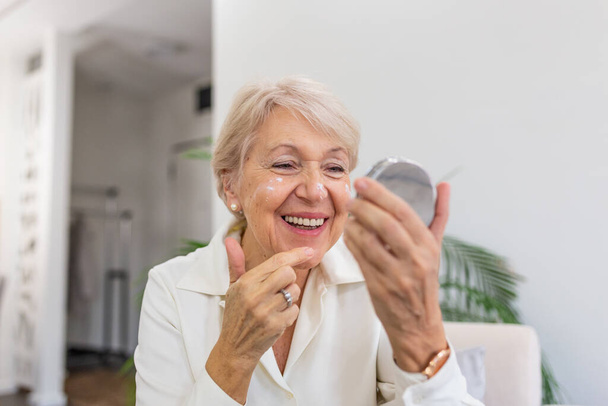 Charming, pretty, old woman touching her perfect soft face skin with fingers, smiling at camera over gray background, using day, night face cream, cosmetology procedures - Φωτογραφία, εικόνα