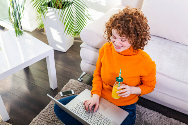 Creative female freelancer sitting front laptop computer and drinking an healthy orange juice. Freelance work Casual red hair woman working on computer while drinking orange juice - Foto, immagini