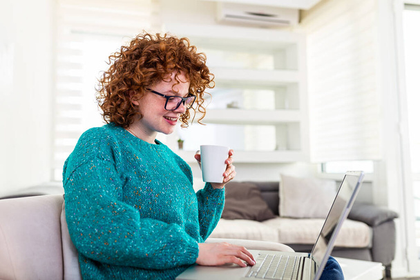 Улыбающаяся рыжая девушка, держащая чашку кофе, работая над ноутбуком у себя дома. Счастливая девочка, отдыхающая на комфортабельном диване и пользующаяся компьютером дома, серфингом в Интернете - Фото, изображение