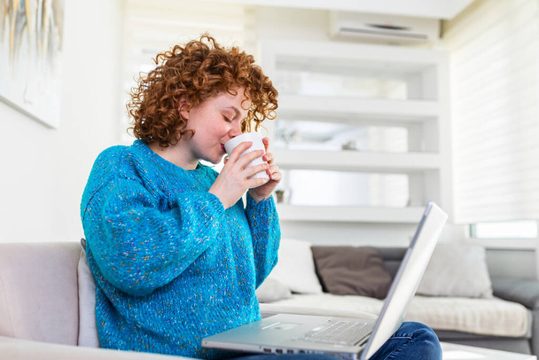 Smiling young redhead holding a cup of coffee while working on her laptop in her home. Happy Girl Relaxing on Comfortable Couch and Using Laptop at Home, Surfing Internet - Foto, afbeelding