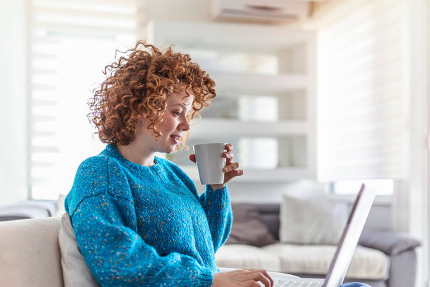 Smiling young redhead holding a cup of coffee while working on her laptop in her home. Happy Girl Relaxing on Comfortable Couch and Using Laptop at Home, Surfing Internet - Fotografie, Obrázek