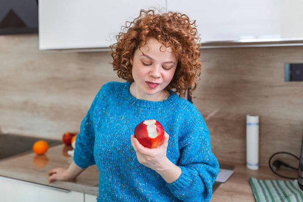 台所でリンゴを噛んだ素敵な笑顔の女の子の肖像画。赤い有機リンゴの実を楽しんでいる若い女性。果物、食事、健康的な食事とビタミンの毎日の摂取量 - 写真・画像