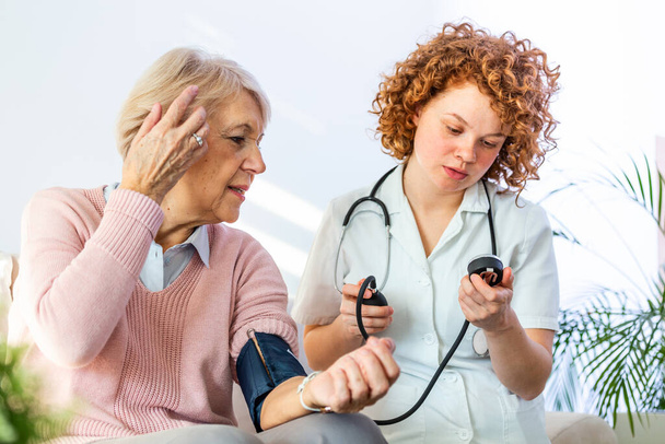Jeune infirmière mesurant la pression artérielle de la femme âgée à la maison. Infirmière vérifiant la pression artérielle d'une femme âgée à la maison, soignante à domicile vérifiant la pression artérielle des patients - Photo, image