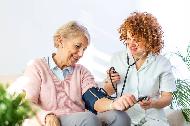 Happy senior woman having her blood pressure measured in a nursing home by her caregiver. Happy nurse measuring blood pressure of a senior woman in living room - Foto, Imagen