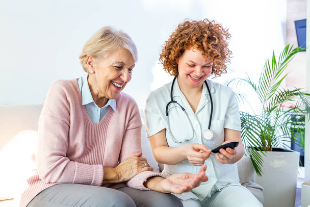 Happy senior woman having her blood sugar measured in a nursing home by her caregiver. Happy nurse measuring blood sugar of a senior woman in living room - diabetes and glicemia concept - Photo, image