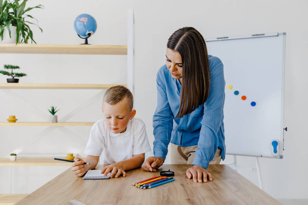 Private lesson. Attentive young woman tutor teacher helping little boy pupil with studying, correct mistakes explain learning material. Smiling mother assist small boy with home task - Foto, afbeelding