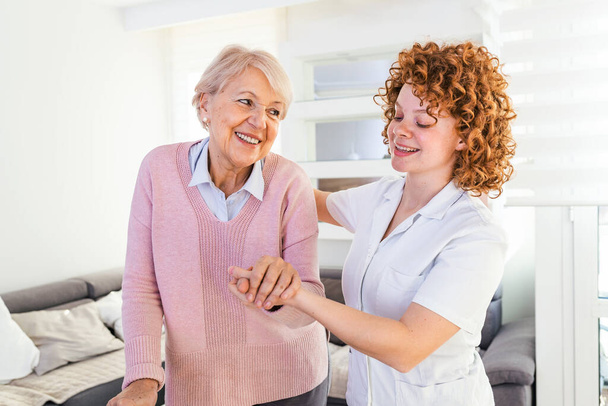 Lächelnde Krankenschwester hilft Seniorin, durch das Pflegeheim zu gehen. Porträt einer glücklichen Pflegerin und einer Seniorin, die gemeinsam zu Hause spazieren gehen. Professionelle Pflegekraft kümmert sich um ältere Frau. - Foto, Bild