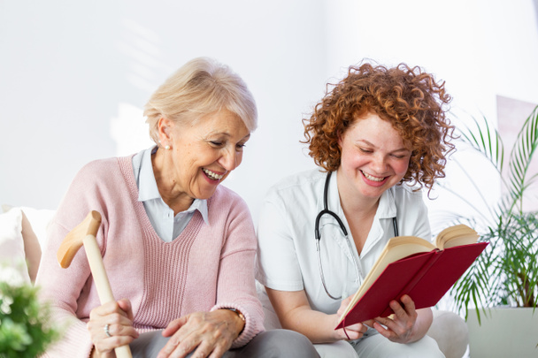 Γυναίκα φροντιστής διαβάζοντας ένα βιβλίο, ενώ κάθεται με ευτυχισμένη ηλικιωμένη γυναίκα στο γηροκομείο. Ευτυχισμένη ηλικιωμένη γυναίκα που κάθεται σε λευκό καναπέ και ακούει τη νοσοκόμα να διαβάζει ένα βιβλίο δυνατά - Φωτογραφία, εικόνα