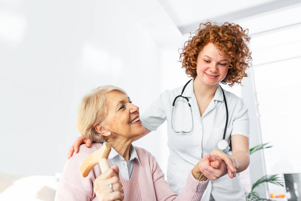 Ευτυχισμένος ασθενής κρατά φροντιστής για ένα χέρι, ενώ περνούν το χρόνο τους μαζί. Ηλικιωμένη γυναίκα στο γηροκομείο και νοσοκόμα. Ηλικιωμένη κομψή γυναίκα στο γηροκομείο - Φωτογραφία, εικόνα