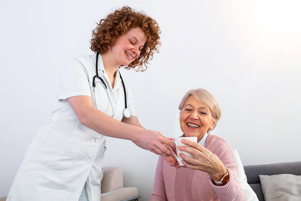 Pretty young caregiver serving afternoon cup of tea to older happy woman. Young nurse caring for elderly patient in her home. Dementia and Occupational Therapy Home caregiver and senior adult woman. - 写真・画像