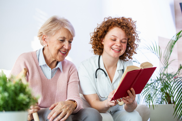 Nainen hoitaja lukee kirjaa istuessaan onnellinen vanhempi nainen hoitokodissa. Onnellinen vanhempi nainen istuu valkoisella sohvalla ja kuuntelee sairaanhoitajaa lukemassa kirjaa ääneen - Valokuva, kuva