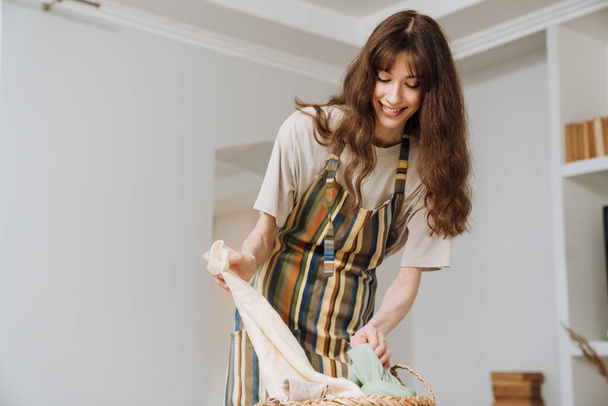 White young woman in apron smiling while doing housework at home - Photo, image