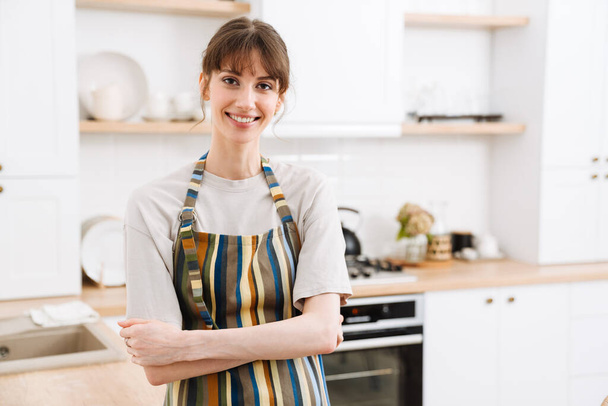 White young woman wearing apron smiling while standing in kitchen at home - Fotó, kép