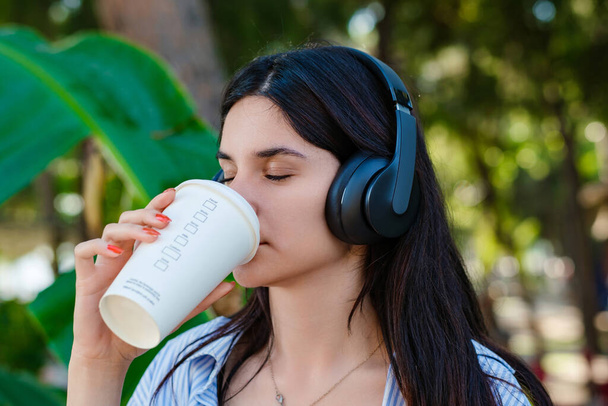 Young caucasian woman wearing shirt standing on city park, outdoors drinking coffee and listening to music. Eyes closed and with cup of coffee in hand, enjoying music, wearing in headphones. - Foto, Imagen