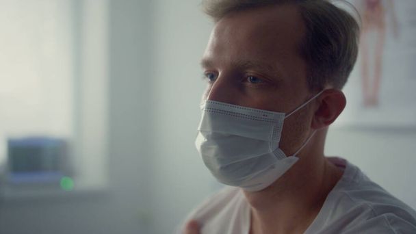 Tired patient sitting wearing mask in modern doctor office closeup. Young serious man holding arm after vaccine injection. Blond guy waiting physician in health clinic. Vaccination healthcare concept. - Foto, immagini