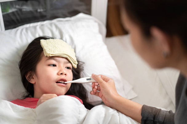 Asian mother measuring temperature girl with digital thermometer in her mouth on bed at morning time, Sick child have cool towel for reduce high fever,  Selective focus, Healthy and infection concept - Фото, зображення