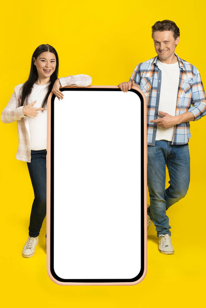 Pointing at white screen caucasian man and asian woman standing leaned on huge smartphone with blank screen, mobile app advertisement isolated on yellow background. Product placement.  - Fotografie, Obrázek