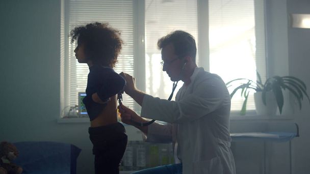 Pediatrician examining teen girl patient lungs using stethoscope in health clinic. African american preteen child breathing deeply standing back to doctor. Young lady visit physician in hospital. - Valokuva, kuva