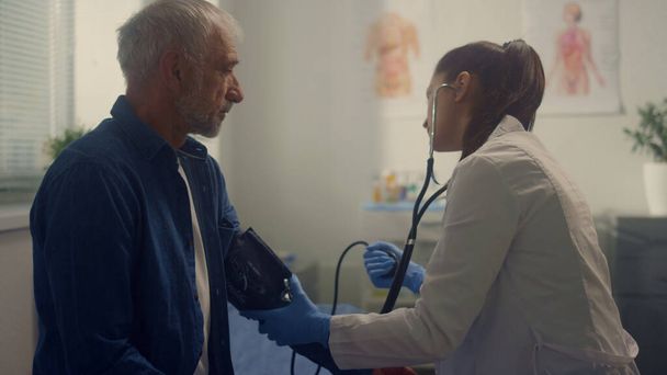 Kardiolog mierzący ciśnienie krwi w gabinecie szpitalnym. Młody lekarz korzystający z tonometru sprawdza stan zdrowia emeryta w nowoczesnej klinice. Koncepcja zapobiegania chorobom.  - Zdjęcie, obraz
