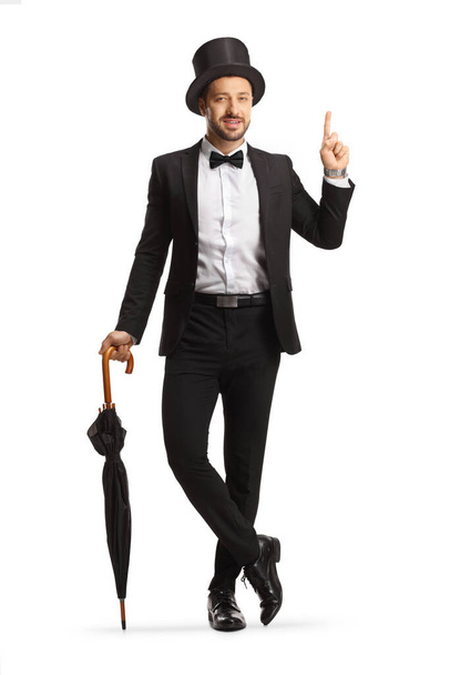 Full length portrait of a young gentleman with a top hat leaning on umbrella and pointing up isolated on white background - Photo, Image