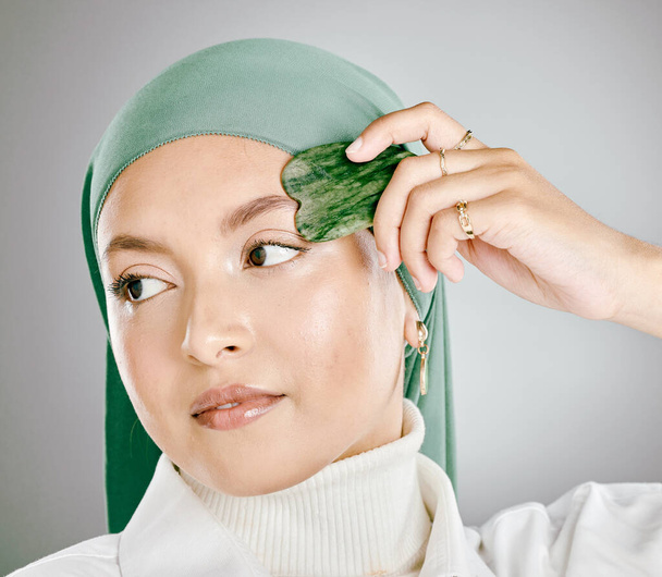 Eine Gua sha, die auf einem jungen muslimischen Frauengesicht verwendet wird, isoliert vor grauem Studiohintergrund. Frau mit Hijab oder Kopftuch, mit einem Anti-Aging-Tool zur Reduzierung von Falten und zur Förderung der Zellerneuerung. - Foto, Bild