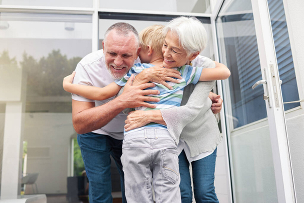 Ältere Großeltern umarmen ihren kleinen Enkel zu Hause. Kleiner Junge umarmt lächelnde Großmutter und Großvater. Adoptiertes Kind fühlt sich glücklich und dankbar, während es älteres Paar umarmt. - Foto, Bild