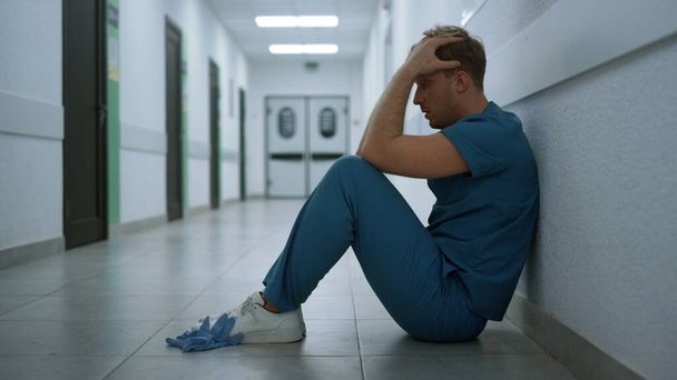 Depressed man surgeon sitting floor clinic hallway close up. Stressed middle age doctor holding head resting in empty corridor side view. Overworking medicine professional after hard pandemic day. - Foto, Imagen
