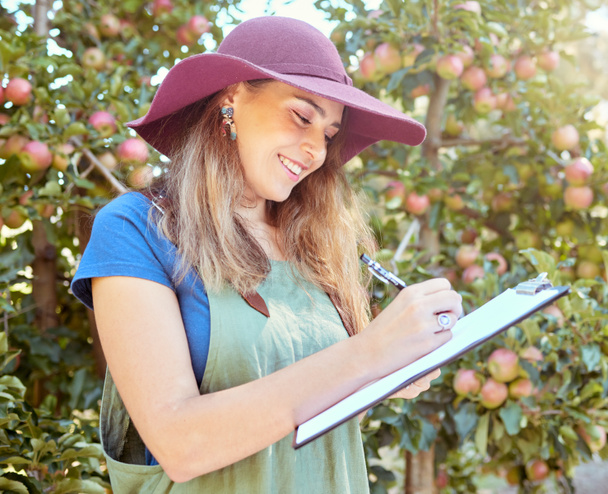 Happy woman writing notes on clipboard while working on sustainable apple orchard farm on sunny day. Cheerful farmer reading checklist while planning task for harvest season of fresh organic fruit. - Foto, afbeelding