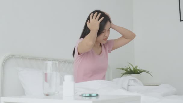 Young asian woman sitting on bed pain headache while drug and water on desk in bedroom at home, unhappy female exhaustion and sick headache having pill treatment, medical and health concept. - Footage, Video