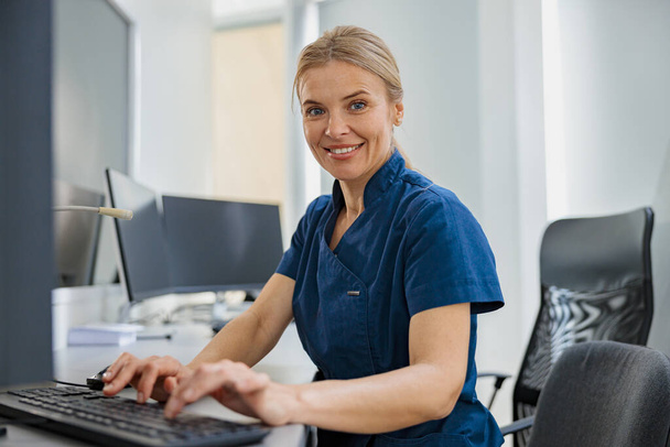 Νοσοκόμα στο καθήκον που εργάζεται στον υπολογιστή στη ρεσεψιόν στη σύγχρονη κλινική. Υψηλής ποιότητας φωτογραφία - Φωτογραφία, εικόνα