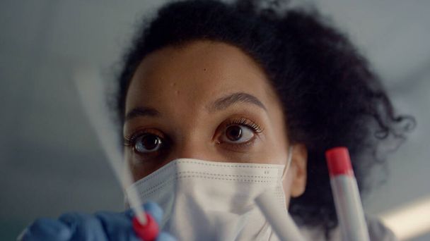 Woman doctor taking sample using cotton swab for coronavirus test close up. African cheerful nurse wearing face mask safety gloves collecting patient saliva in clinic. Young physician holding tube. - 写真・画像