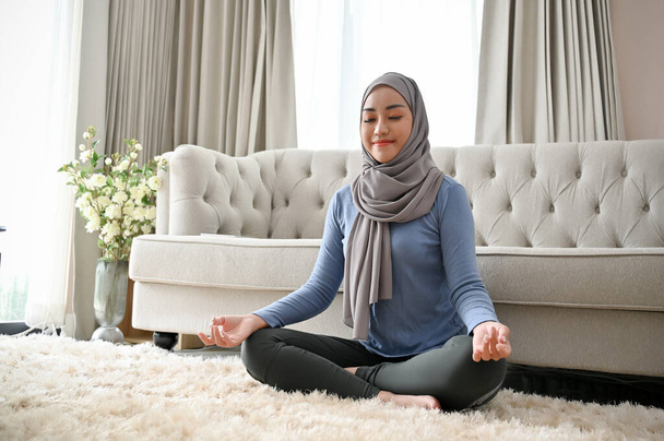 Atrakcyjna, piękna, tysiącletnia azjatycka muzułmanka z hidżabem lub chustą ćwiczącą jogę, skoncentrowana medytacja, pozująca lotosem w salonie. - Zdjęcie, obraz