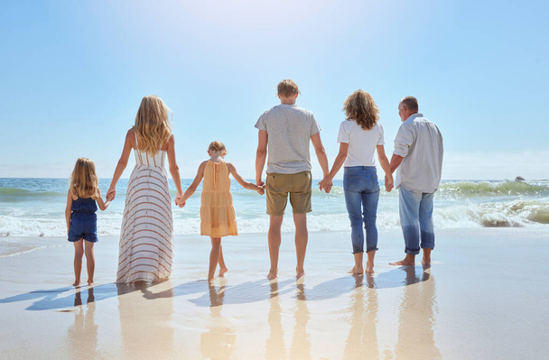 Rear view of family holding hands and having fun on a beach vacation together on a sunny day. Relatives enjoying summer and sunshine, bonding and spending quality time walking and feeling ocean water. - Zdjęcie, obraz