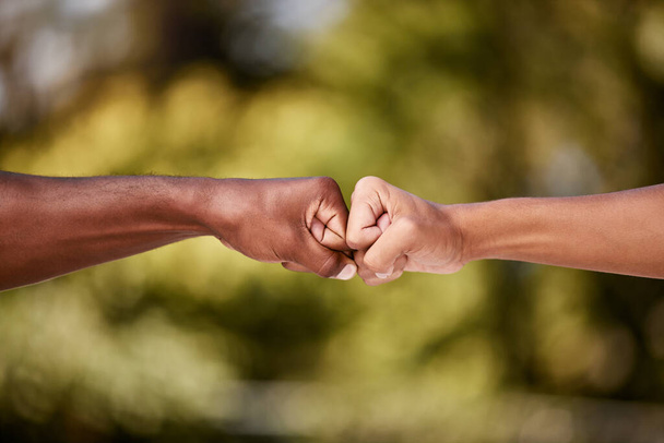 Fist bump of two interracial men outdoor against a blur background. Closeup of diverse athletes doing social gesture greeting in a park. Showing solidarity, friendship, brotherhood, teamwork or unity. - Photo, image