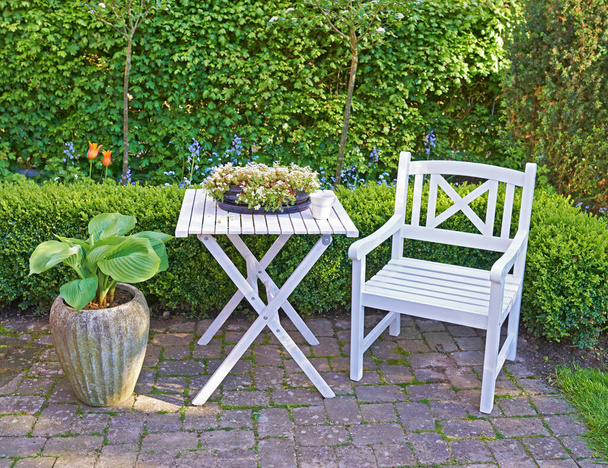 White wooden chair and table in a green garden with blossoming plants and copyspace. Tranquil landscape to relax and enjoy a cosy picnic in summertime. Outdoor patio in a quiet and secluded courtyard. - Foto, imagen