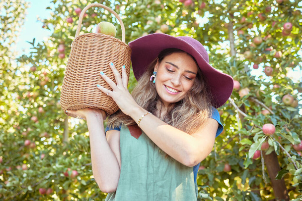 Jonge lachende vrouw met een emmer vol appels. Een vrouwtje met een zak vol biologisch fruit in een boomgaard tijdens het oogstseizoen buiten. Boeren die fruit oogsten van bomen op een boerderij. - Foto, afbeelding